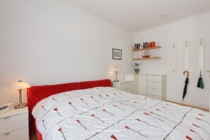 Un dormitorio con una cama, una mesita de noche, una lámpara, un armario, una estantería con libros y una planta, y un paraguas colgado en la pared.