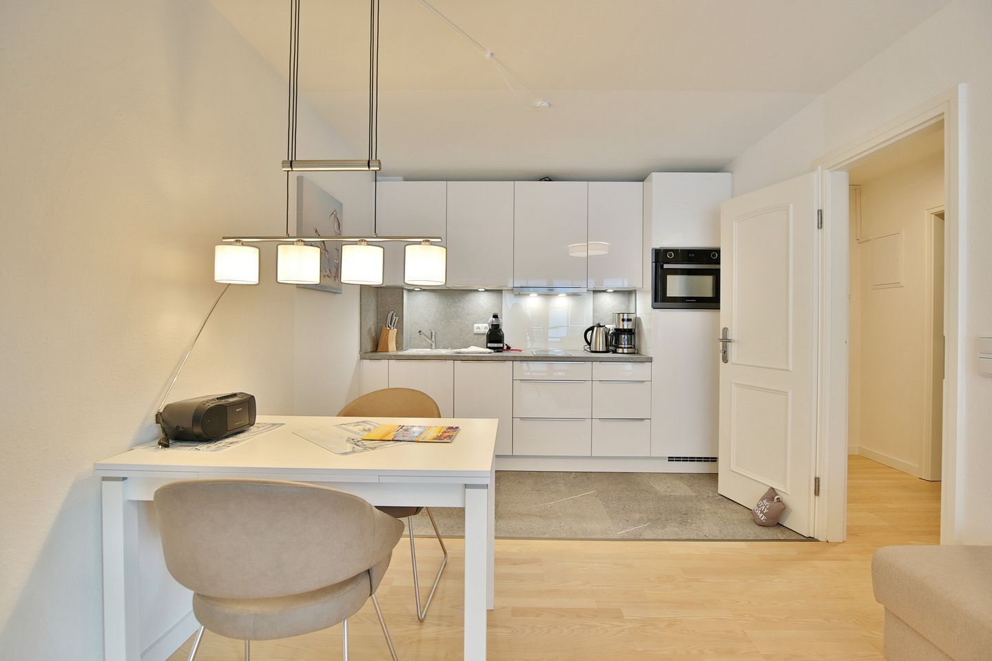 A compact kitchen with white cabinets, a coffee machine, and a microwave. There is a dining table with a chair and a lamp.