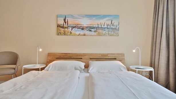 A hotel room with two beds, a wooden headboard, two bedside tables with lamps, a chair, and a painting of a beach scene on the wall. - Seerose II Apartment 14 - House Seerose (Scharbeutz)