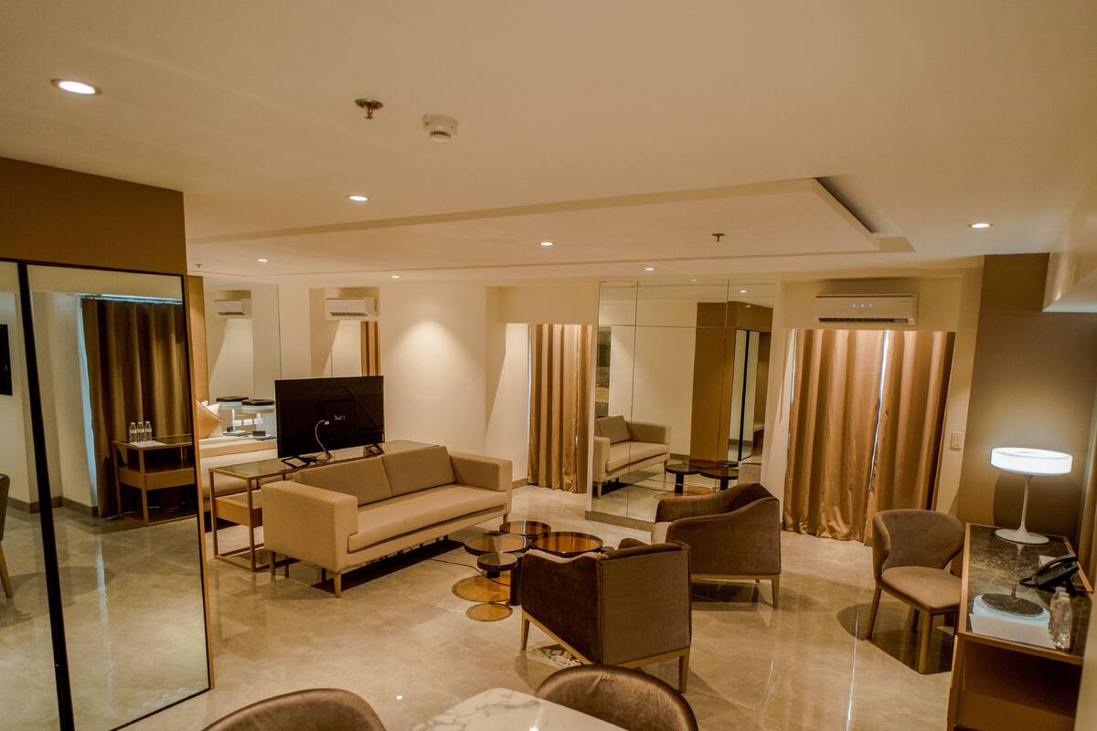 Una habitación de hotel moderna con sofá, sillones, televisor y espejo.