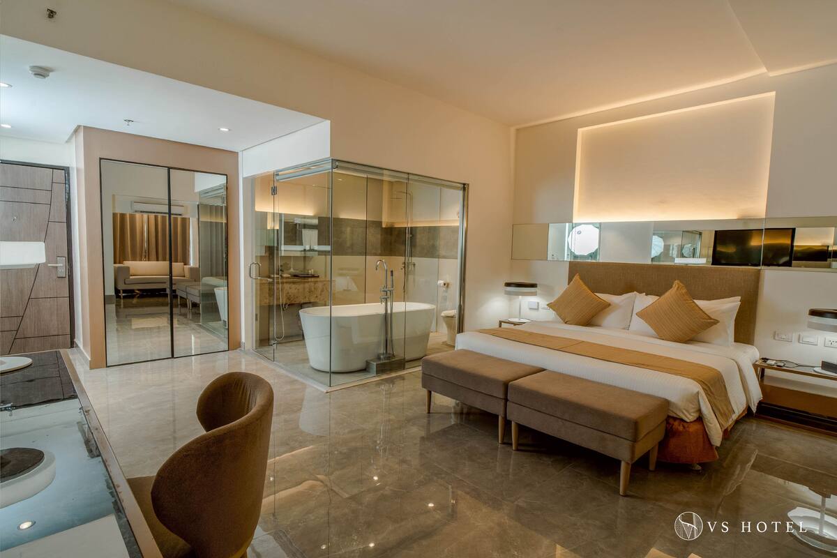 Una habitación de hotel moderna con una cama grande, una bañera, zona de ducha con mampara de vidrio y un área de descanso con una silla y un banco.