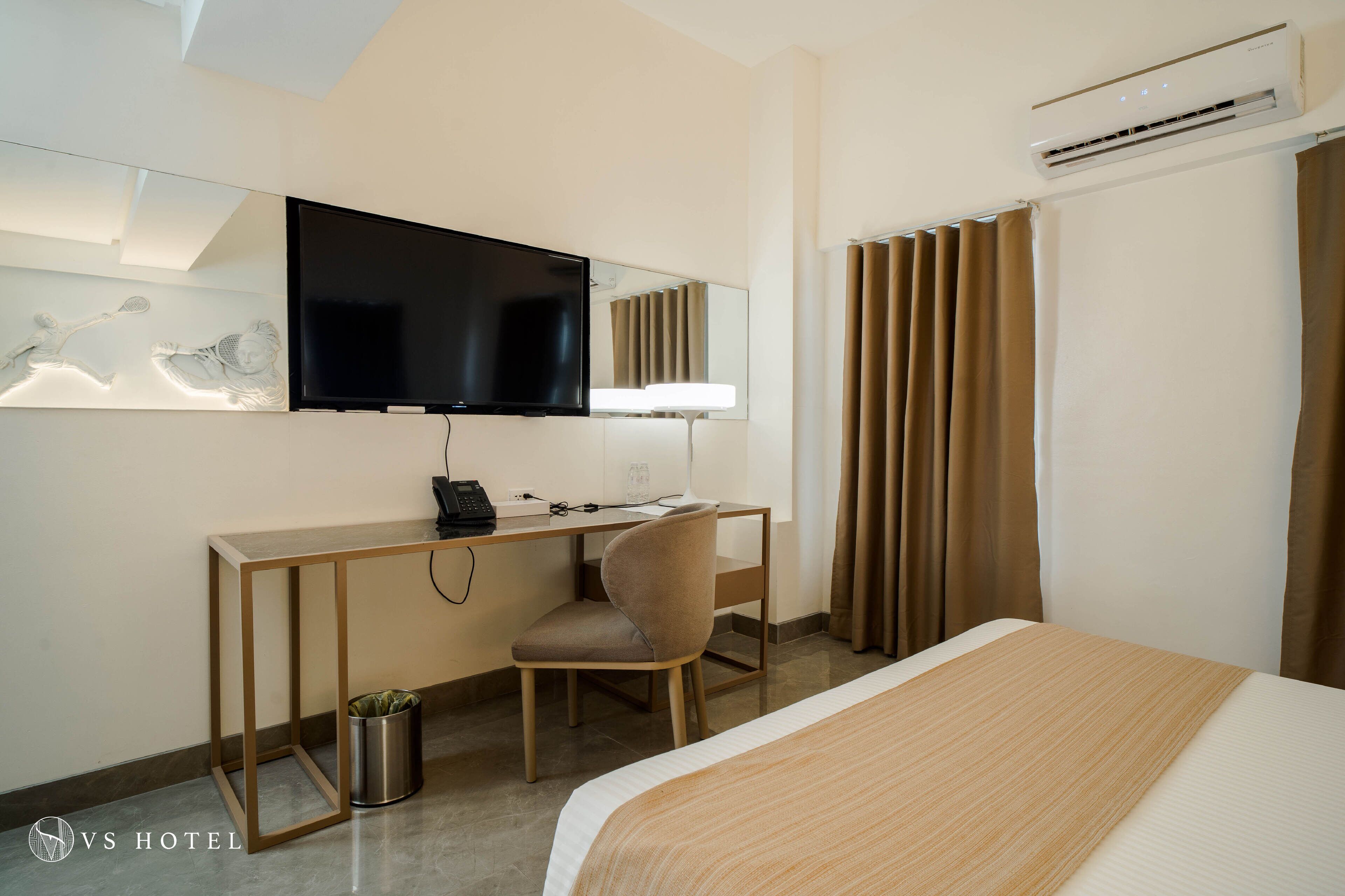 A hotel room with a bed, a desk with a chair, a television, and an air conditioning unit.