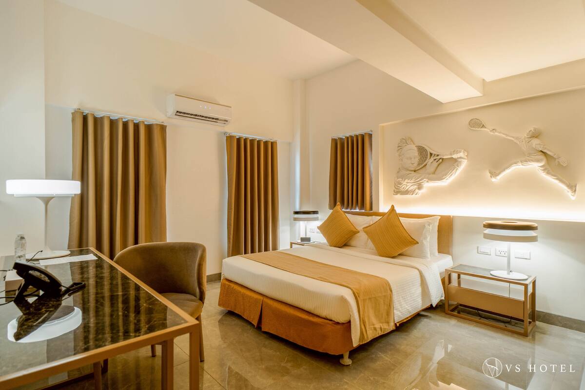 Habitación de hotel con cama, escritorio, silla, mesita de noche y lámpara de pared.
