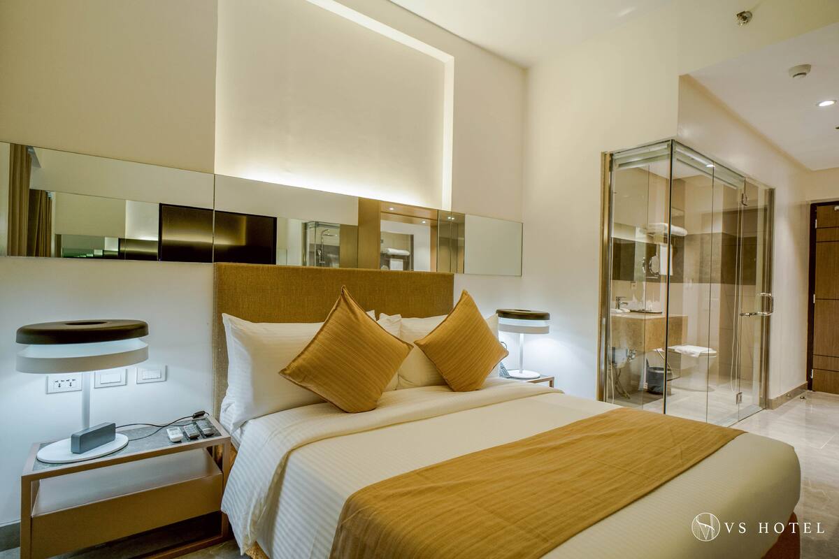 Habitación de hotel moderna con una cama grande, una mesita de noche con lámpara, un bidet con teléfono y cargador, y un área de ducha con paredes de vidrio.