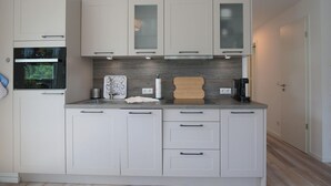 Una cocina moderna con armarios blancos, horno integrado, cafetera y tabla de cortar de madera.