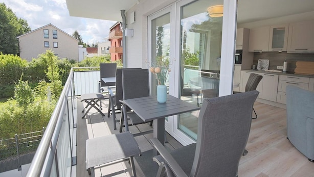 Un balcón con mesa y sillas, una puerta de cristal que da a una cocina, y vistas de edificios y vegetación circundantes.