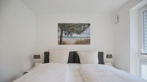 Un dormitorio con una cama, dos mesitas de noche, una lámpara de pared y un cuadro con una escena de playa.