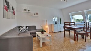A modern living room with a grey sofa, a white coffee table, and a dining area with wooden chairs and a table. There is a white cabinet with a mirror above it, and a potted plant on the coffee table. - Domizil Seestraße - Ihr Domizil Seestraße (Scharbeutz)