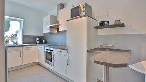 A modern kitchen with white cabinets, a built-in oven, a microwave, and a coffee maker. There is a small shelf with a plant and a bowl. - Domizil Seestraße - Ihr Domizil Seestraße (Scharbeutz)