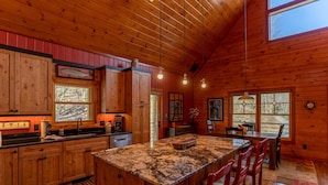 A wooden cabin interior with a kitchen featuring a granite island, wooden cabinets, and a dining area with red chairs. - A River Mist Walk - Walk to river, pet friendly, hot tub, pool table (Sugar Grove)