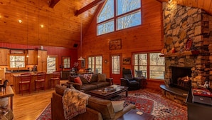 A spacious log cabin interior with a stone fireplace, large windows, and a cozy living area. - A River Mist Walk - Walk to river, pet friendly, hot tub, pool table (Sugar Grove)