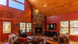 A cozy cabin interior with a stone fireplace, wooden walls, and large windows offering views of the outdoors. - A River Mist Walk - Walk to river, pet friendly, hot tub, pool table (Sugar Grove)