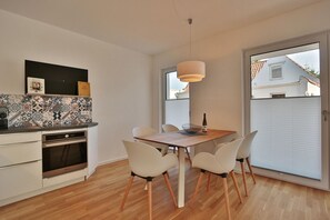 A modern kitchen with white cabinets, a built-in oven, and a patterned backsplash. There is a dining table with white chairs and a hanging light fixture above it.