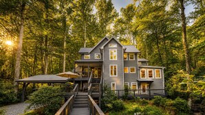 Une maison à plusieurs étages dotée d’une terrasse donnant sur un étang, le tout entouré d’une forêt dense.