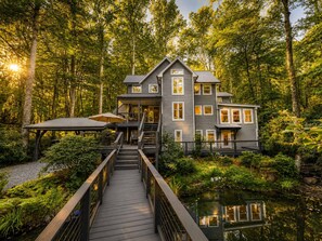 A multi-story house with a deck leading to a pond, surrounded by dense forest.