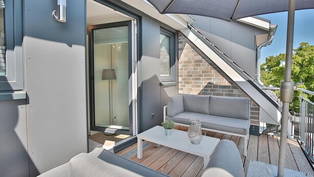 A modern outdoor deck with a sofa, table, and umbrella, featuring a glass door leading to a room with a lamp. - Bude 37 Apartment 6 - Bude 37 (Scharbeutz)