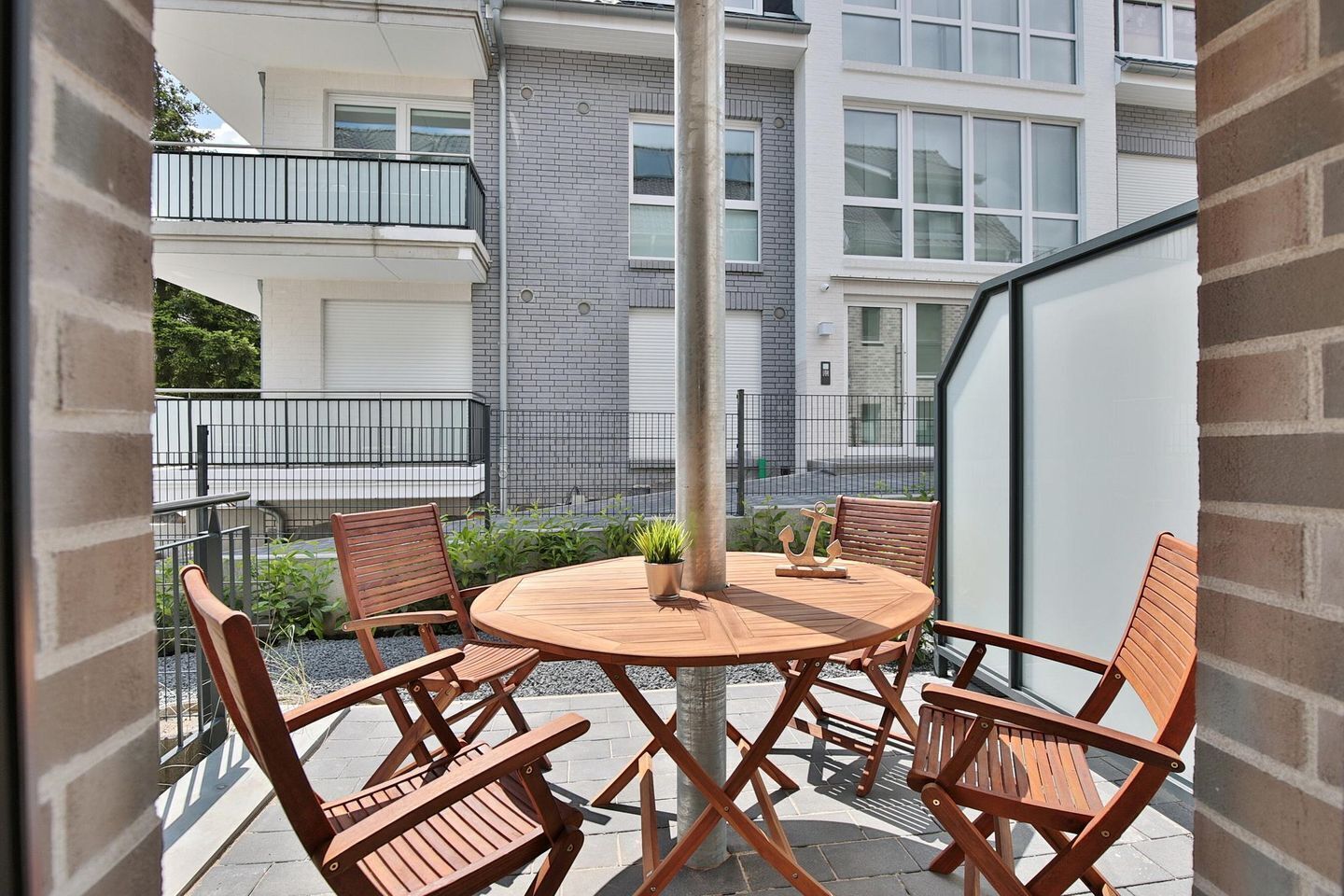 A balcony with a round wooden table and four wooden chairs, surrounded by a brick wall and a building with balconies.