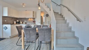 Une cuisine moderne avec des armoires blanches, un plan de travail en bois et un coin repas comprenant deux chaises grises et une table. Un escalier avec une rampe en métal mène à l’étage supérieur.