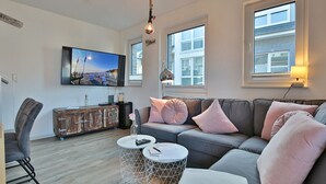 Un salon moderne avec un canapé gris, une malle ancienne, un téléviseur à écran plat et une grande fenêtre donnant sur la ville.
