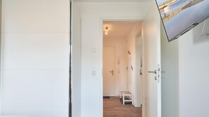 Une chambre d’hôtel avec un lit, un plancher en bois, une armoire blanche, une porte donnant sur un couloir et un téléviseur fixé au mur.