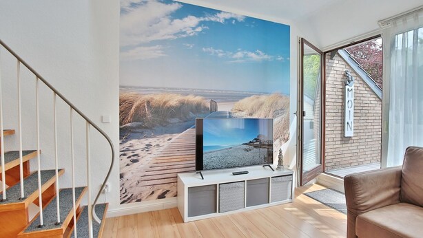 A modern living room with a large TV on a white stand, a wooden staircase, and a view of the beach through a large window. - Haus Adria Appartement 31 - Haus Adria (Scharbeutz)