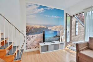 A modern living room with a large TV on a white stand, a wooden staircase, and a view of the beach through a large window. - Haus Adria Appartement 31 - Haus Adria (Scharbeutz)