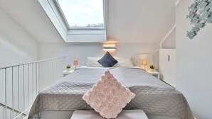 A bedroom with a skylight, a bed with a quilted headboard, bedside lamps, and a decorative wall piece. - Haus Adria Appartement 31 - Haus Adria (Scharbeutz)
