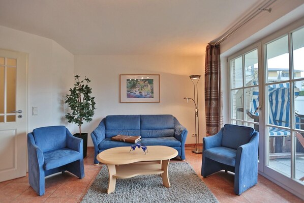 Ein Wohnzimmer mit einem blauen Sofa, zwei blauen Sesseln, einem Holztisch, einer Stehlampe, einem Blumentopf und einem großen Fenster mit Vorhängen.