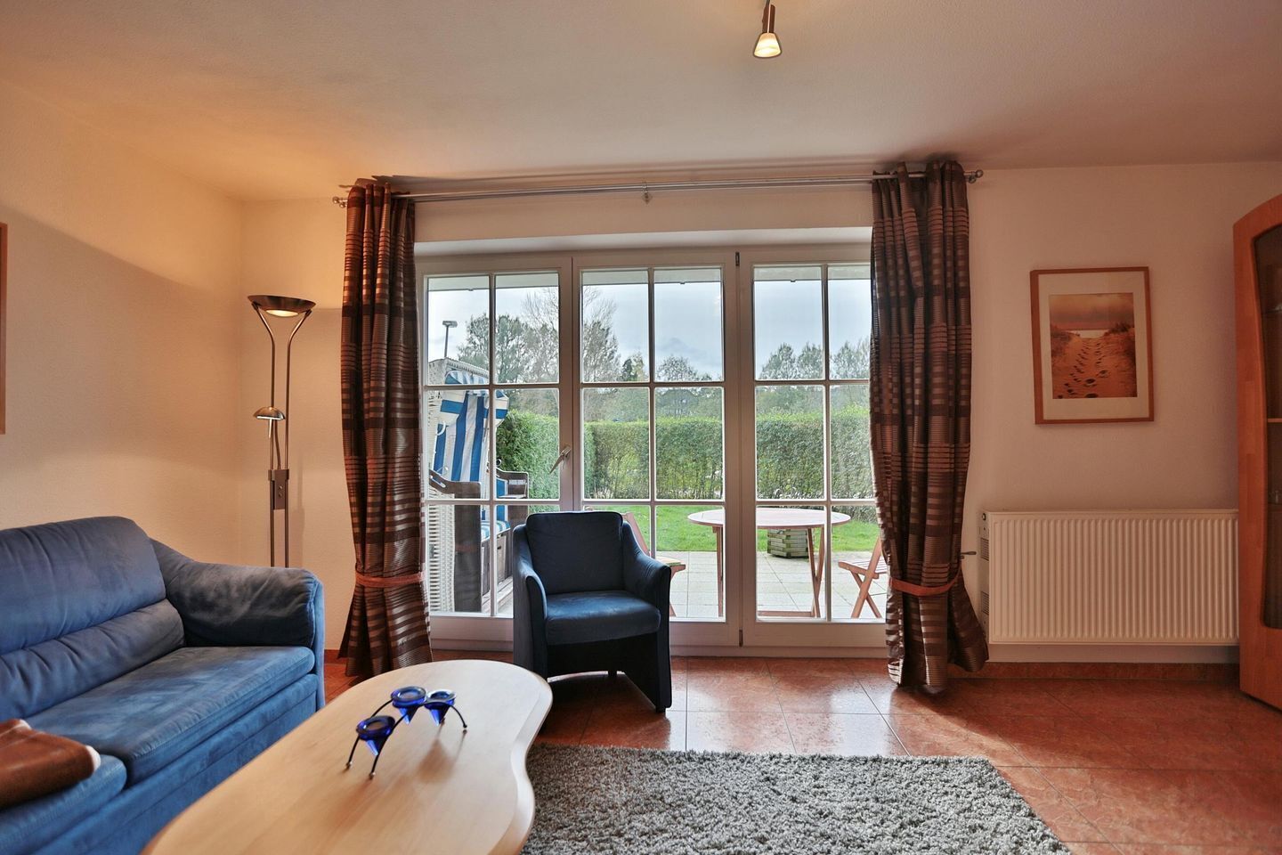 A living room with a blue sofa, a black chair, a wooden coffee table, and a view of a garden through large windows.