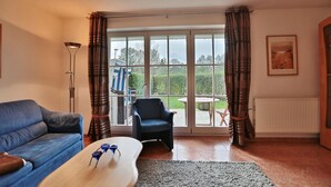Ein Wohnzimmer mit einem blauen Sofa, einem schwarzen Sessel, einem Holztisch und Blick auf einen Garten durch große Fenster.