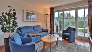 Ein Wohnzimmer mit einem blauen Sofa, einem Holztisch und einem blauen Sessel. Es gibt ein großes Fenster mit Vorhängen, einen Topfpflanzen und ein gerahmtes Bild an der Wand.