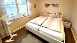 A bedroom with a large bed, a window with blinds, a bedside table with a lamp, and a painting on the wall.