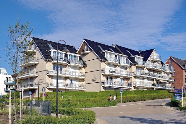 A row of modern multi-story residential buildings with balconies, a paved driveway, and a clear sky.