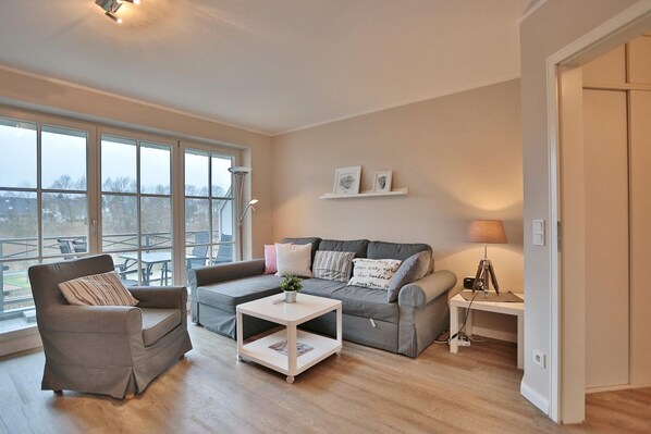 A modern living room with a grey sofa, a brown armchair, and a white coffee table. There are large windows providing a view of the outdoors.
