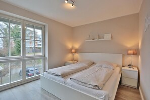 A bedroom with a large window, two bedside tables with lamps, a bed with white bedding, and a shelf above the bed. - Hanseaten Residence Apartment 15 - Hanseaten Residence (Scharbeutz)