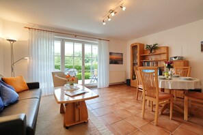 Ein Wohnzimmer mit Sofa, einem Esstisch mit Stühlen, einem Bücherregal und einem Fenster mit Vorhängen.