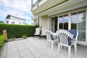 An outdoor patio with a table and chairs, a hedge, and a building with a balcony.