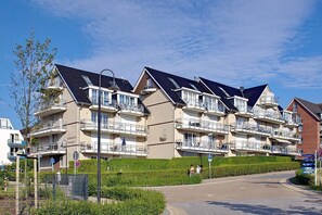 A row of modern multi-story residential buildings with balconies, a paved driveway, and a clear sky.
