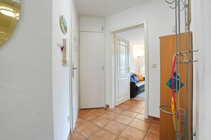Ein Flur mit Fliesenboden, einer Tür zu einem Zimmer mit Bett und Lampe sowie einem Kleiderständer mit Gegenständen.