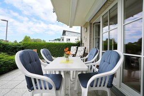 A covered outdoor patio with a white table and chairs, a potted plant, and a view of a modern building.