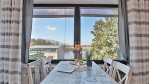 A dining table with a nautical-themed tablecloth, white chairs, and a view of a lake and buildings through large windows.