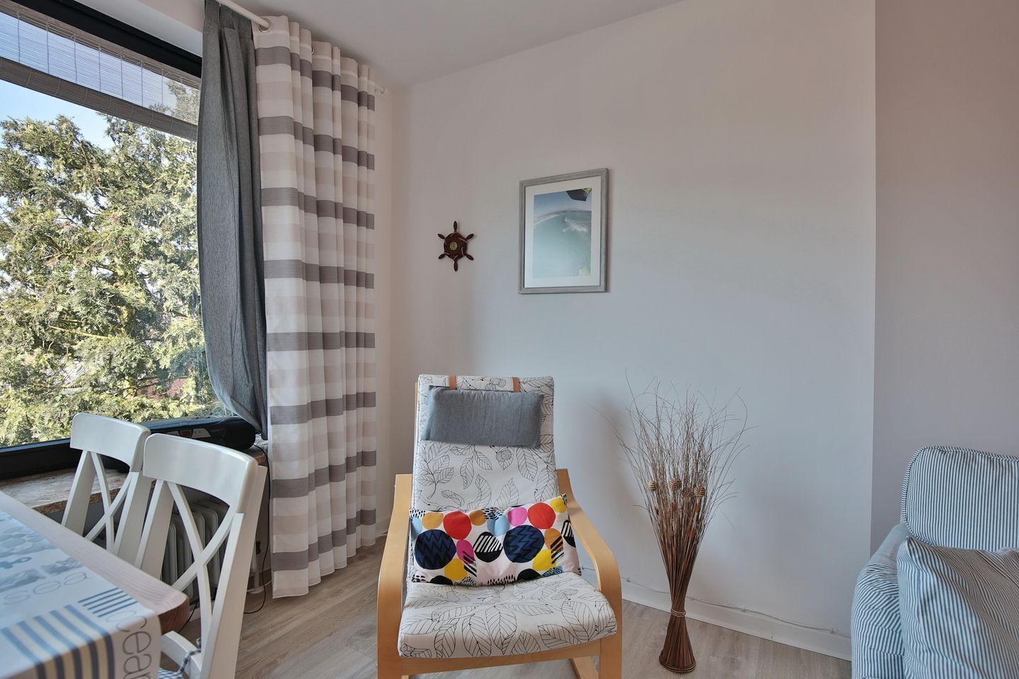 Ein gemütliches Zimmer mit einem Holzstuhl, einem Esstisch und Blick auf Bäume durch ein Fenster mit gestreiften Vorhängen.