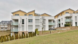 Un edificio residencial moderno con varios balcones, techo plano y un área verde con árboles jóvenes.