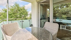 Ein Balkon mit Korbstühlen, einem Glastisch und einer Schiebetür, die zu einem Poolbereich führt, mit Blick auf Bäume und Gebäude.