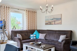 A living room with a brown sofa, a coffee table, a painting of birds on a beach, and a chandelier. - Dünenhaus 3 Appartement 7 - Dünenhaus (Scharbeutz)