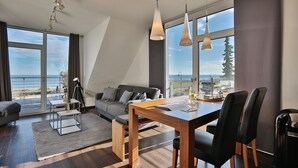 A modern living room with a sofa, dining table, and chairs. Large windows offer a view of the outdoors.