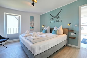 A modern bedroom with a bed, towels, a sailboat painting, a wall-mounted lamp, and a window.