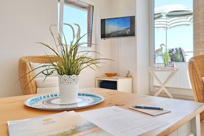 A room with a wooden table, a potted plant, a television, and a view of the beach.