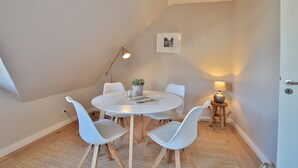 Una habitación pequeña y acogedora con una mesa redonda, cuatro sillas blancas, una pequeña planta en maceta, una lámpara de pie y un cuadro en la pared.