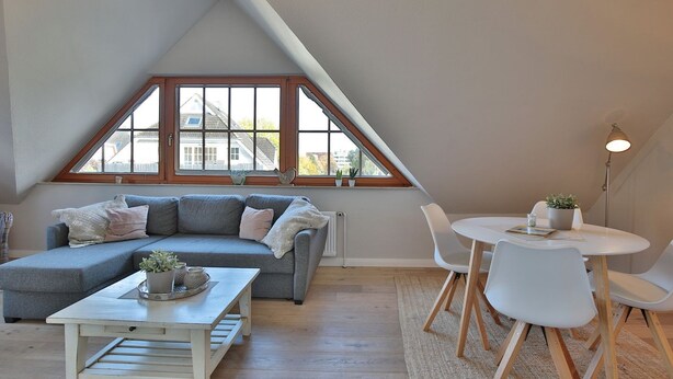 Una habitación estilo loft con un sofá seccional gris, una pequeña mesa blanca y una mesa de comedor redonda con sillas. Un ventanal ofrece vistas al exterior.